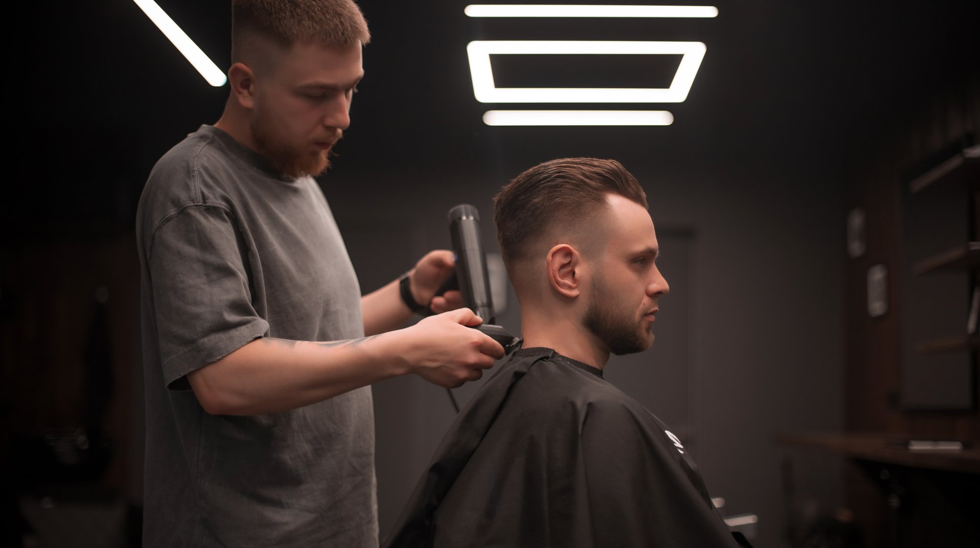 A male barber styles a client's hair using a hairdryer in a modern, dimly lit barbershop. The client sits still, covered with a black cape, during the final touch of his grooming session.